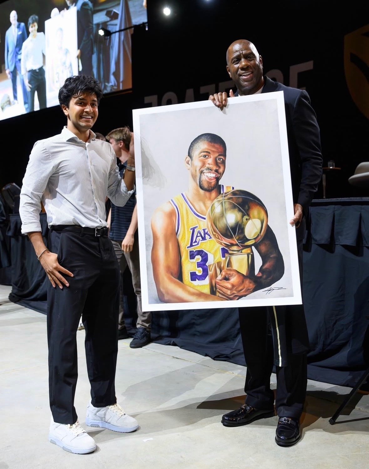 Magic Johnson on stage holding a Kyan Patel portrait of a Lakers player holding the NBA championship trophy